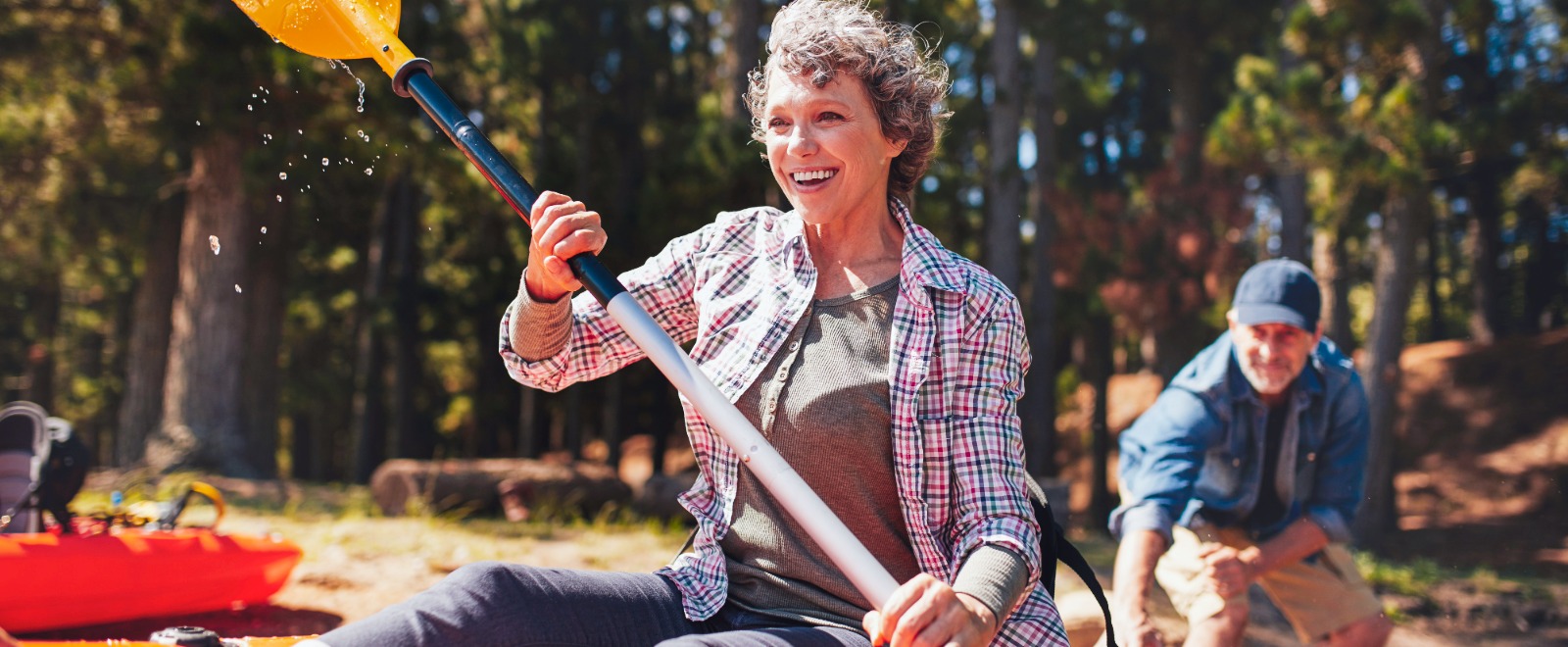 Retired woman paddles in a kayak while a retired man watches from the bank.